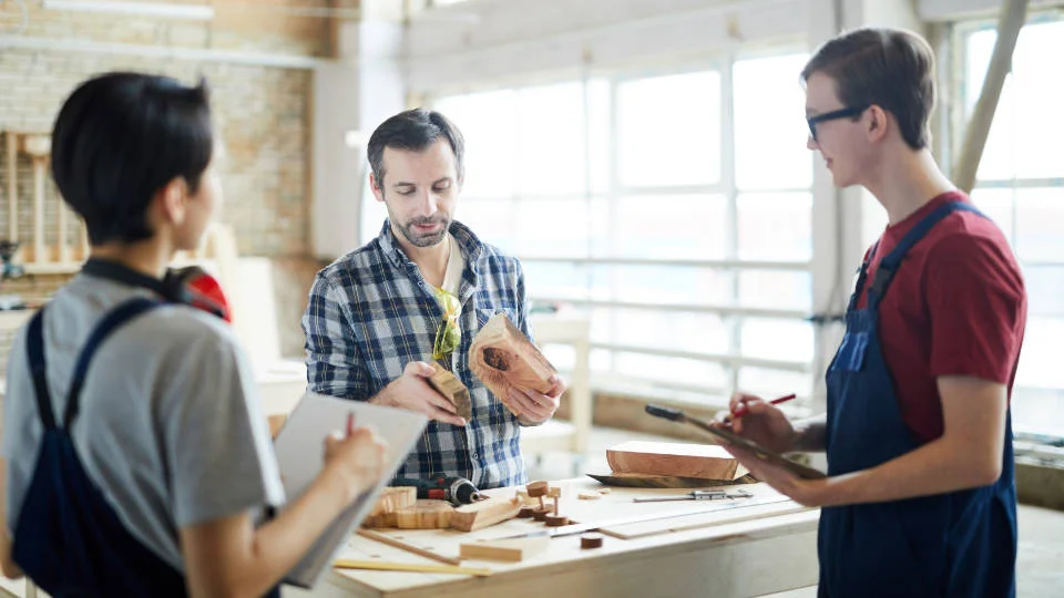 Adult skills in the Nordic region–abstract Three people in a woodworking workshop with one person measuring a wooden piece, another taking notes, and a third observing.