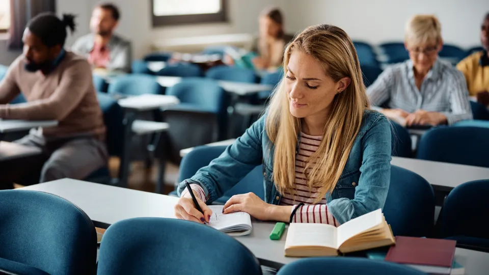 Finland satsar på framtidens kompetens för alla. Kvinna skriver anteckningar i ett klassrum med andra studenter i bakgrunden.