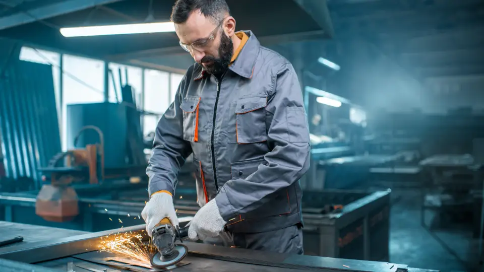 Kompetanse kan ikke lades i løpet av natta – langsiktig og kontinuerlig innsats må til. Arbetare som använder en vinkelslip i en metallverkstad, iförd skyddskläder och skyddsglasögon.
