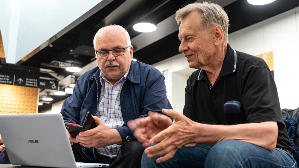Seniorer i Finland får hjälp med att använda digitala verktyg To ældre mænd sidder og arbejder sammen på en bærbar computer, hvoraf den ene holder en smartphone.