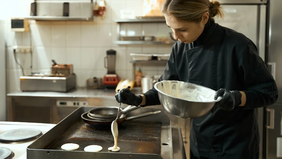 En kock klädd i svart arbetsklädsel häller smet på en stekhäll för att tillaga pannkakor eller plättar. Köksmiljön är välutrustad och ren med professionella redskap och maskiner i bakgrunden.