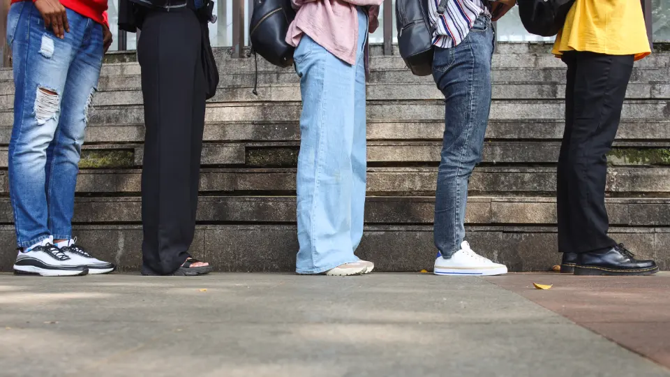 Trods høj beskæftigelse er 43.000 danske unge stadig uden job og uddannelse Billedet viser en gruppe personer stående i kø. Fokus er på deres ben og fødder, som er klædt i forskelligt tøj og fodtøj. Trinene i baggrunden antyder en offentlig eller udendørs placering, og personerne har rygsække, hvilket kan tyde på en hverdagssituation som venten på transport eller adgang til en bygning.