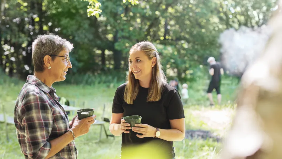 New one-pager on how to become a teacher that promotes sustainability within adult education To kvinder står i en skovlysning og snakker, mens de holder kopper. Solen skinner gennem træerne, og i baggrunden ses flere personer, herunder et barn og en mand i sort tøj.