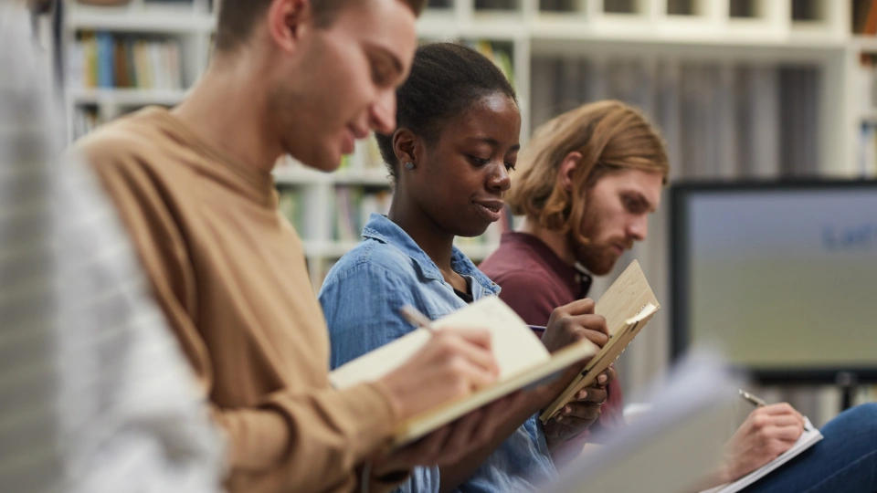 Yrkesprofiler identifierar grundläggande färdigheter för utbildare Tre unga vuxna sitter i ett bibliotek och skriver i anteckningsböcker med fokus.