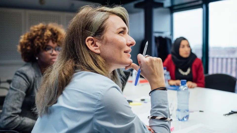 Regeringen uppmanar till stöd för kvinnors företagande Kvinnor i möte runt ett konferensbord, en av dem håller en penna och lyssnar uppmärksamt.