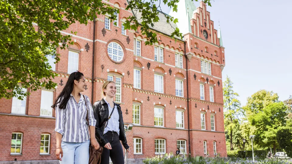 En utredning tillsätts om att stärka den akademiska friheten Två unga kvinnor promenerar framför en historisk röd tegelbyggnad med gröna tak i en lummig parkmiljö.