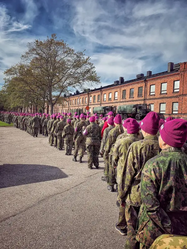 En lång rad personer i militäruniform och lila mössor marscherar utomhus vid en kasern.
