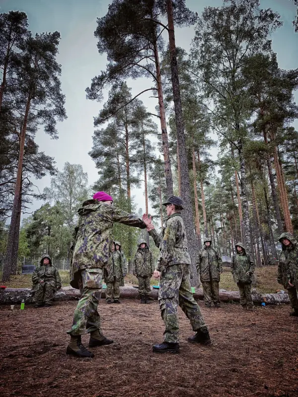 Två personer i militäruniform tränar självförsvar i en skog med andra deltagare i bakgrunden.
