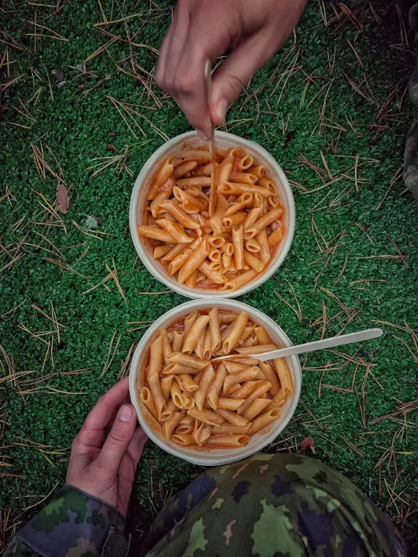 Två skålar med pennepasta hålls av personer i militäruniform, en hand rör om med plastgaffel.