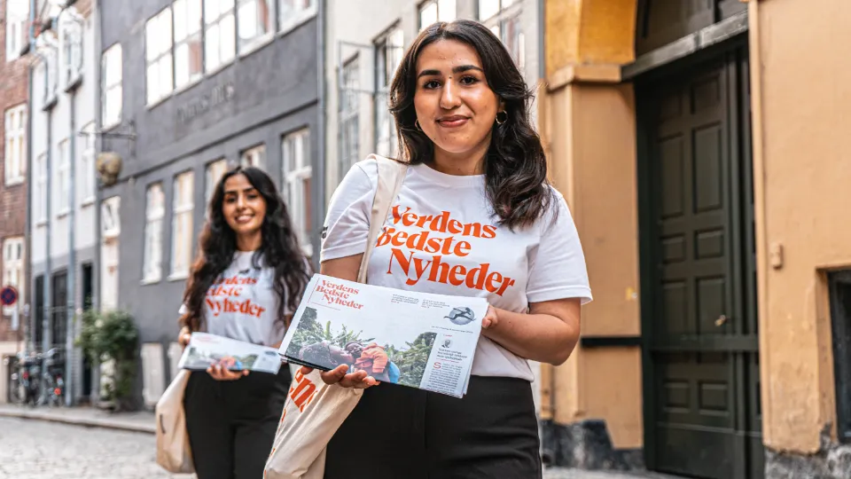 Verdens Bedste Nyheder skaber balance i mediebilledet To kvinder iført t-shirts med teksten "Verdens Bedste Nyheder" uddeler aviser på en brostensbelagt gade i en historisk bydel.