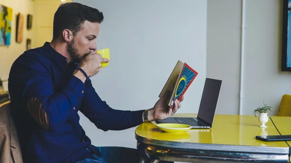 Man som sitter vid ett gult bord med en bärbar dator, dricker kaffe och läser en bok i en modern inomhusmiljö.