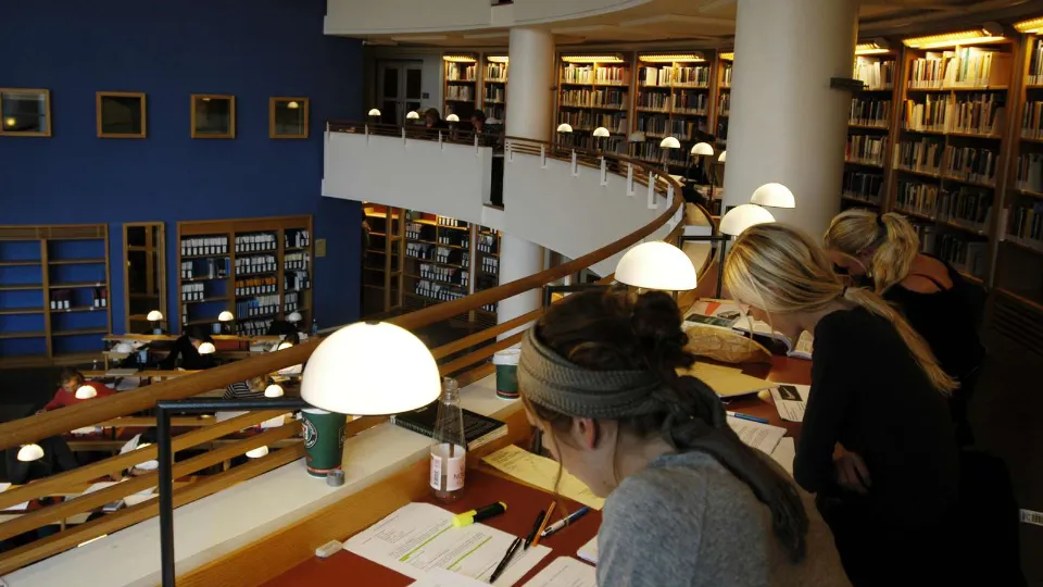 Studenter studerar vid skrivbord i ett bibliotek med böcker och läslampor.