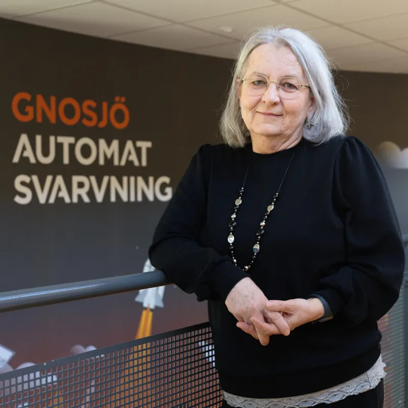 Äldre kvinna inomhus, Elvira Zahirovic. Bakom henne vägg där det står Gnosjö automatsvarvning.