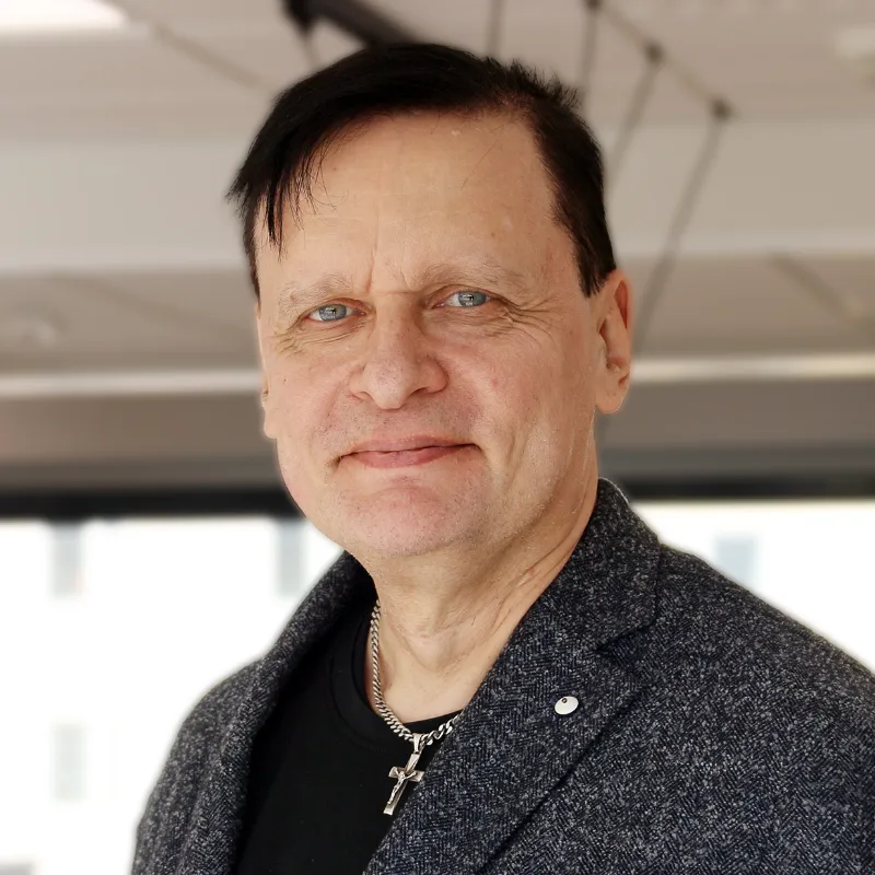 Heikki Tulkki med kort mörkt hår, klädd i mörk kavaj och korshalsband, fotograferad inomhus med ljus bakgrund.
