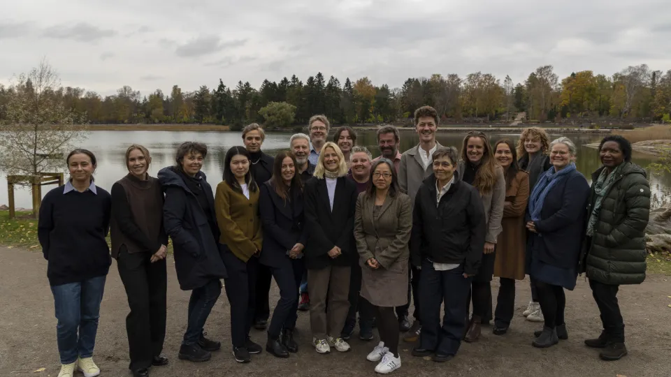 Group photo of diverse adults standing outdoors by a lake in autumn, posing together and smiling.