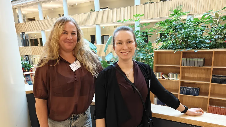 Victoria Nylund og Linda Heinola Victoria Nylund og Linda Heinola smilende foran en bogreol i et lyst og moderne kontormiljø med grønne planter i baggrunden.