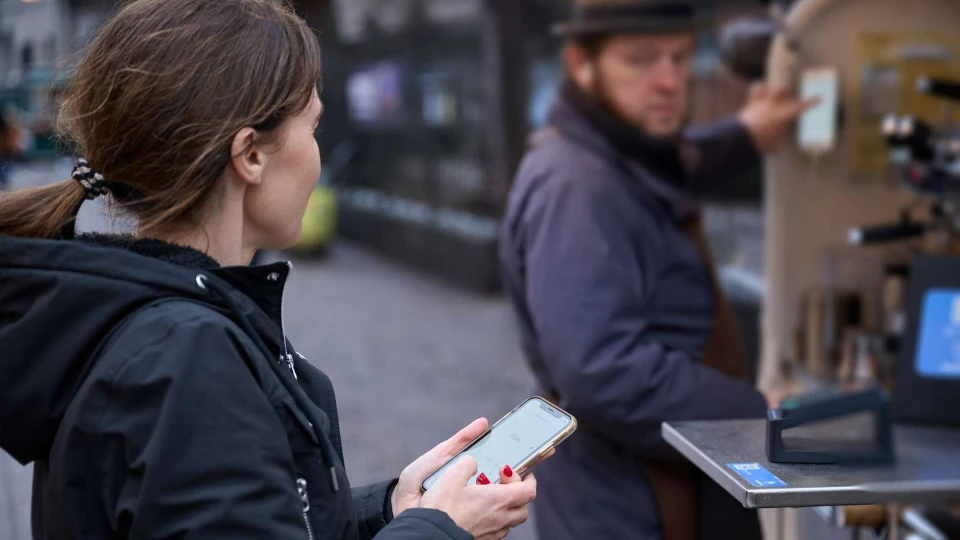 Digital integration Kvinde betaler med mobiltelefon ved en udendørs kaffevogn, hvor en barista forbereder en bestilling.