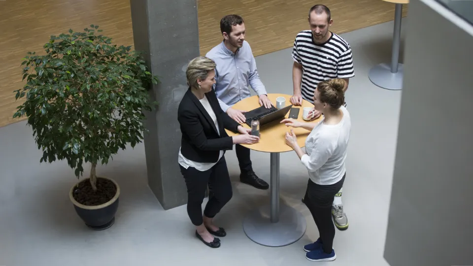 Fire personer står omkring et højt bord i et kontormiljø og diskuterer foran en bærbar computer.