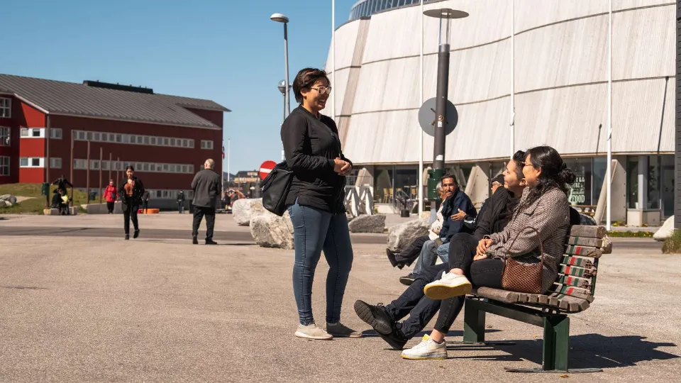 Grønland kickstarter Gruppe mennesker taler og slapper af på en bænk i solrigt bymiljø med moderne og traditionelle bygninger i baggrunden.