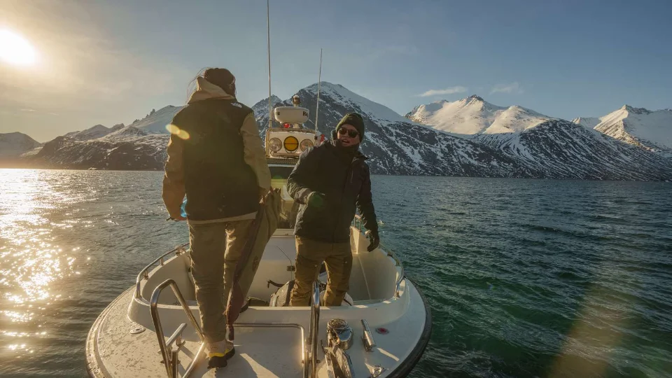 To mænd på en båd i en fjord omgivet af snedækkede bjerge under en lav vintersol.