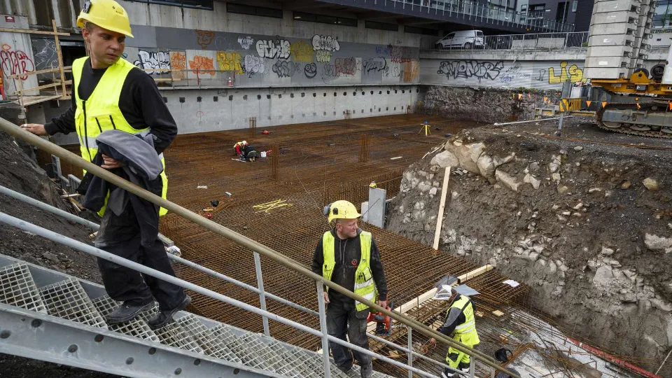 Byggeplads med håndværkere i gang med at lægge armeringsjern til et fundament i en dyb udgravning.