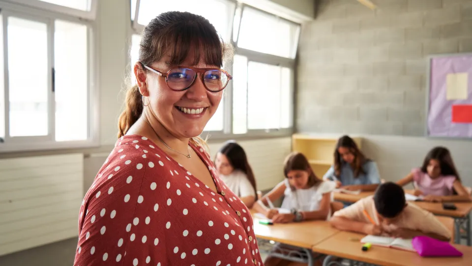 En smilende kvindelig lærer med briller og prikket bluse står i et klasseværelse, mens elever arbejder koncentreret ved deres borde i baggrunden.
