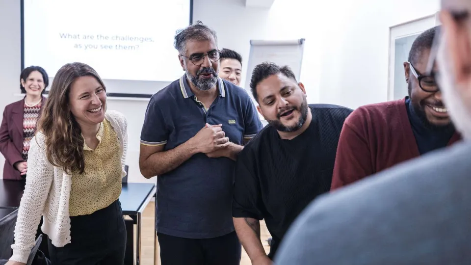 Smilende kolleger i et mødelokale under workshop, hvor en gruppe diskuterer foran en projektorskærm.