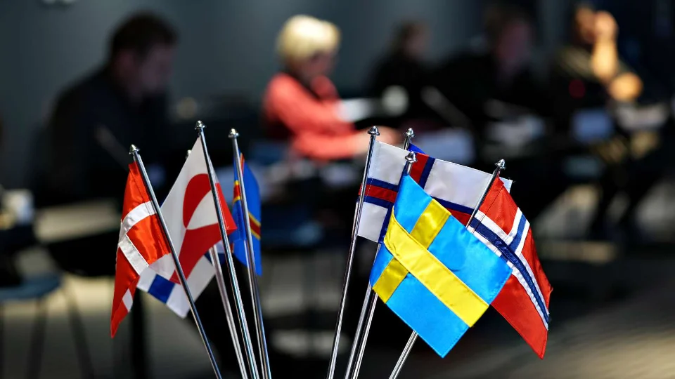 Small Nordic desk flags (Denmark, Greenland, Faroe Islands, Sweden and Norway) in the foreground with blurred people in a meeting room background.