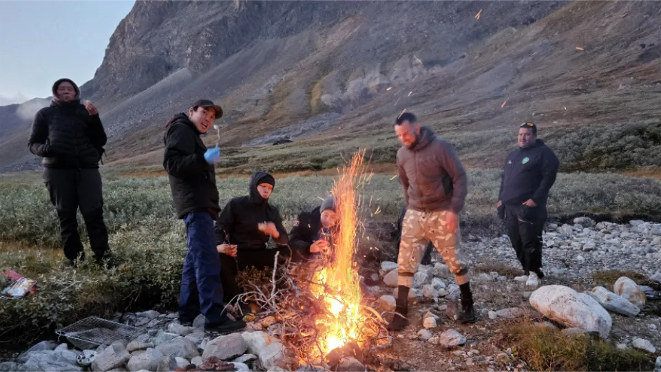 Gruppe af mennesker samles omkring et bål i et bjerglandskab ved skumring.