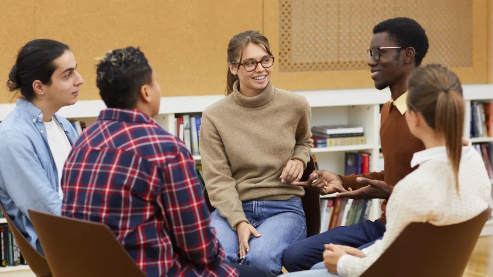 Fem personer sitter i en cirkel och samtalar i ett bibliotek eller klassrum.