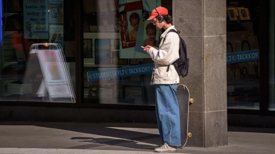 Person med röd keps, hörlurar och ryggsäck står med skateboard utanför en butik och tittar på sin mobil.