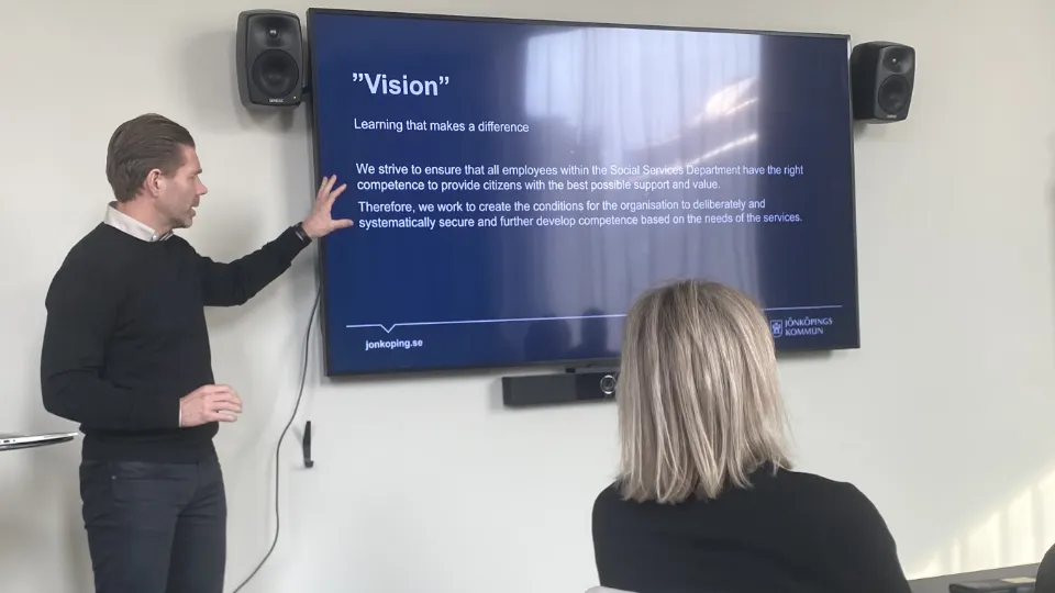 En mann holder en presentasjon foran en stor skjerm med teksten «Vision», mens en person sitter og ser på i et møterom.