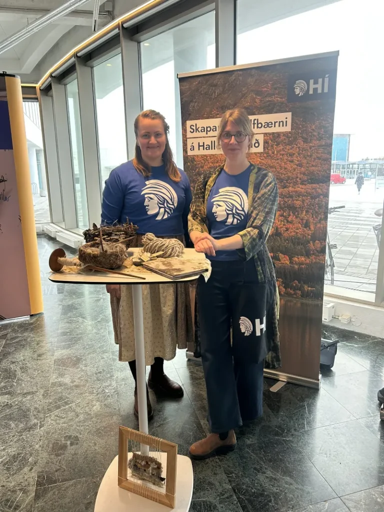 To personer, Myrra Mjöll Daðadóttir (til venstre), står ved et bord med håndverksmaterialer foran en HI-rollup.