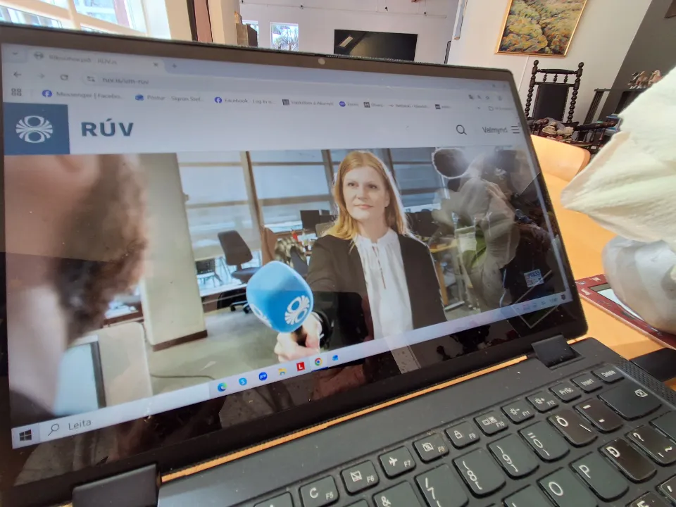 Bærbar PC som viser RÚVs nettside med et intervju på skjermen, fotografert på et bord i et rom med stoler og kunst på veggene.