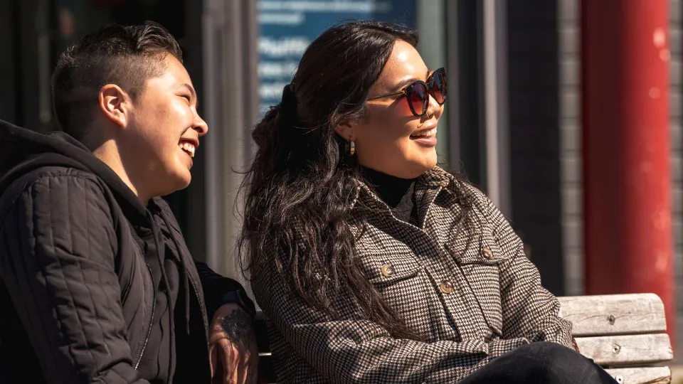 Two smiling people sitting outdoors on a bench in bright sunlight in an urban setting.