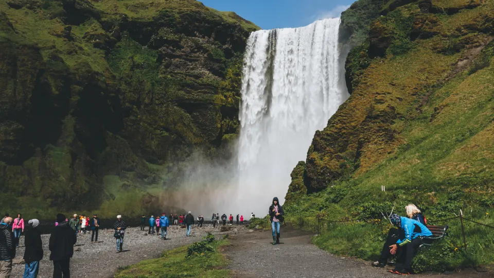 En satsning på utbildning inom turism Ett stort vattenfall på Island faller ner från en grön klippvägg, medan turister står och går på stigen framför och beundrar utsikten.