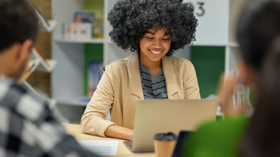 En smilende person med krøllet hår sidder ved en bærbar computer ved et bord, omgivet af andre slørede personer i et studie- eller arbejdsmiljø.