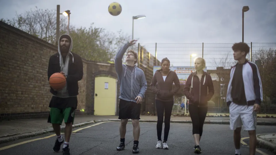 Fem unge voksne går på en urban gate med basketball og fotball ved en inngjerdet bane.