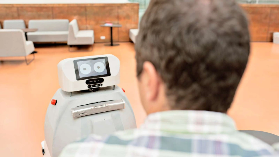 Framtidens kompetensbehov är svårare att förutse än någonsin En person sitter med ryggen mot kameran och tittar på en liten robot med skärmögon i en ljus inomhusmiljö.