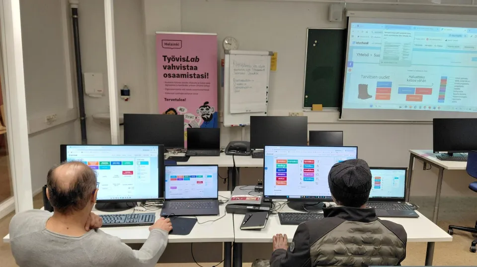 Two people working at computers in a classroom with a projector, whiteboard, and screens for digital learning.